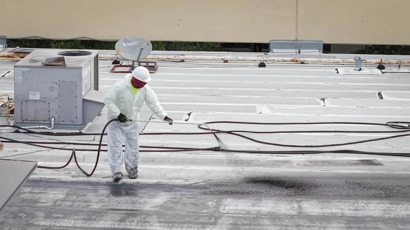 A worker in a white protective suit, hard hat and face covering spraying a low-slope roof a few feet from a packaged rooftop unit, with a hose running across the roof