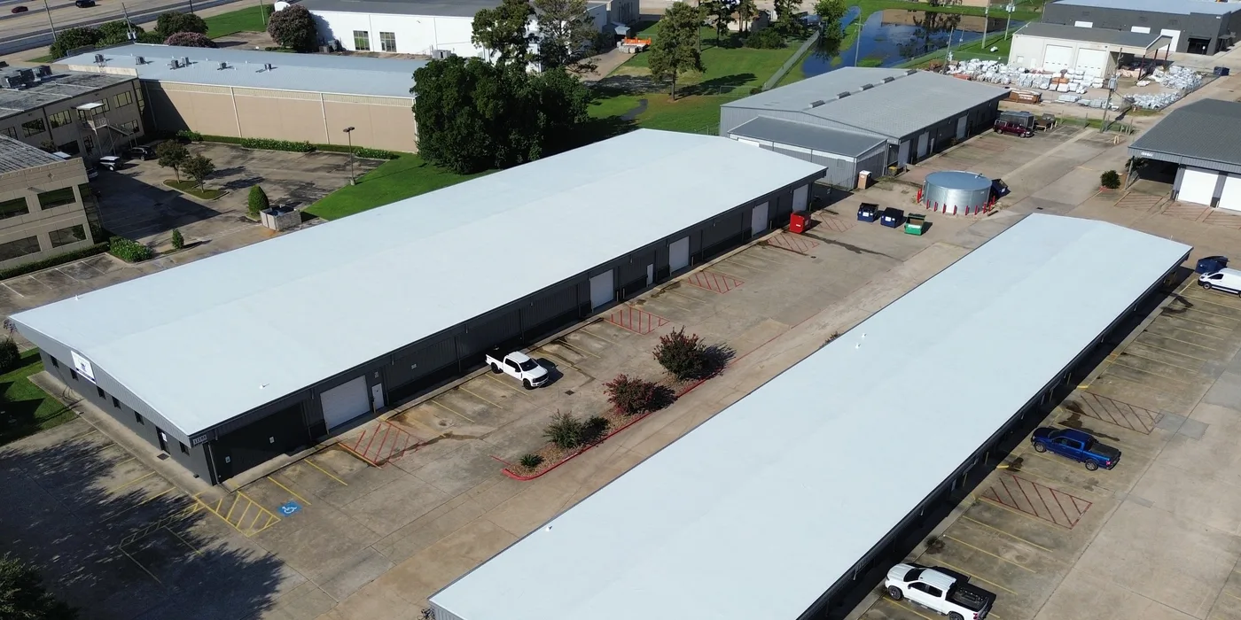 Aerial view of two long single-story metal buildings with white roofs and roll-up doors along their sides, a parking lot between them and older gray metal buildings beyond
