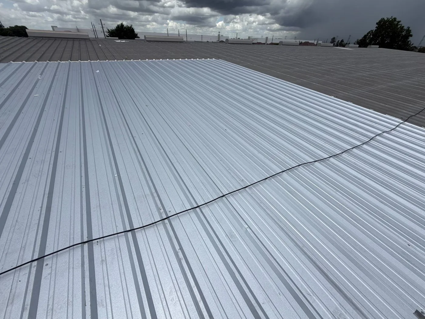 A large section of bright new ribbed metal panels on an older gray-brown metal roof, under a dark storm sky, with a black cable lying across the new panels
