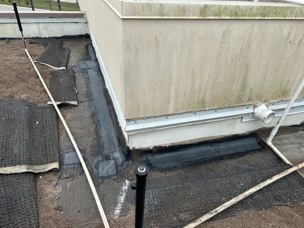 Base flashing along a stuccoed wall repaired in black reinforced membrane that carries on around the corner, with walkway pads and white pipes nearby