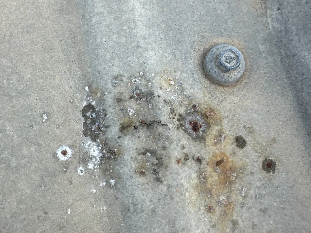 Close-up of a weathered gray metal roof panel with a cluster of rust pits, white residue rings around some of them, a short hairline crack and a fastener nearby