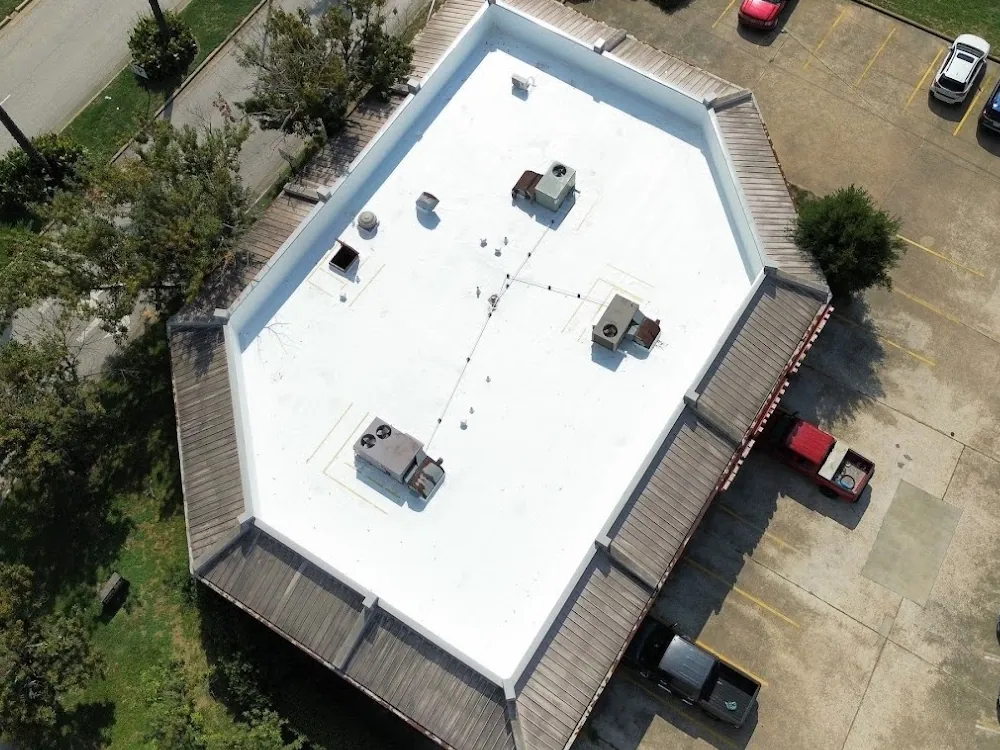 Aerial view of a white low-slope commercial roof inside a mansard edge, with rooftop units, vents and a pipe run across it, and parked cars beside the building