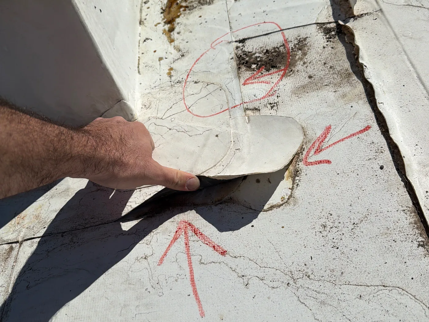 A hand lifting a loose, rounded patch off a weathered white TPO roof at the base of a flashed corner, with red marker arrows pointing at the lifted patch and a red circle around a stained, cracked area where the fabric inside the sheet shows through