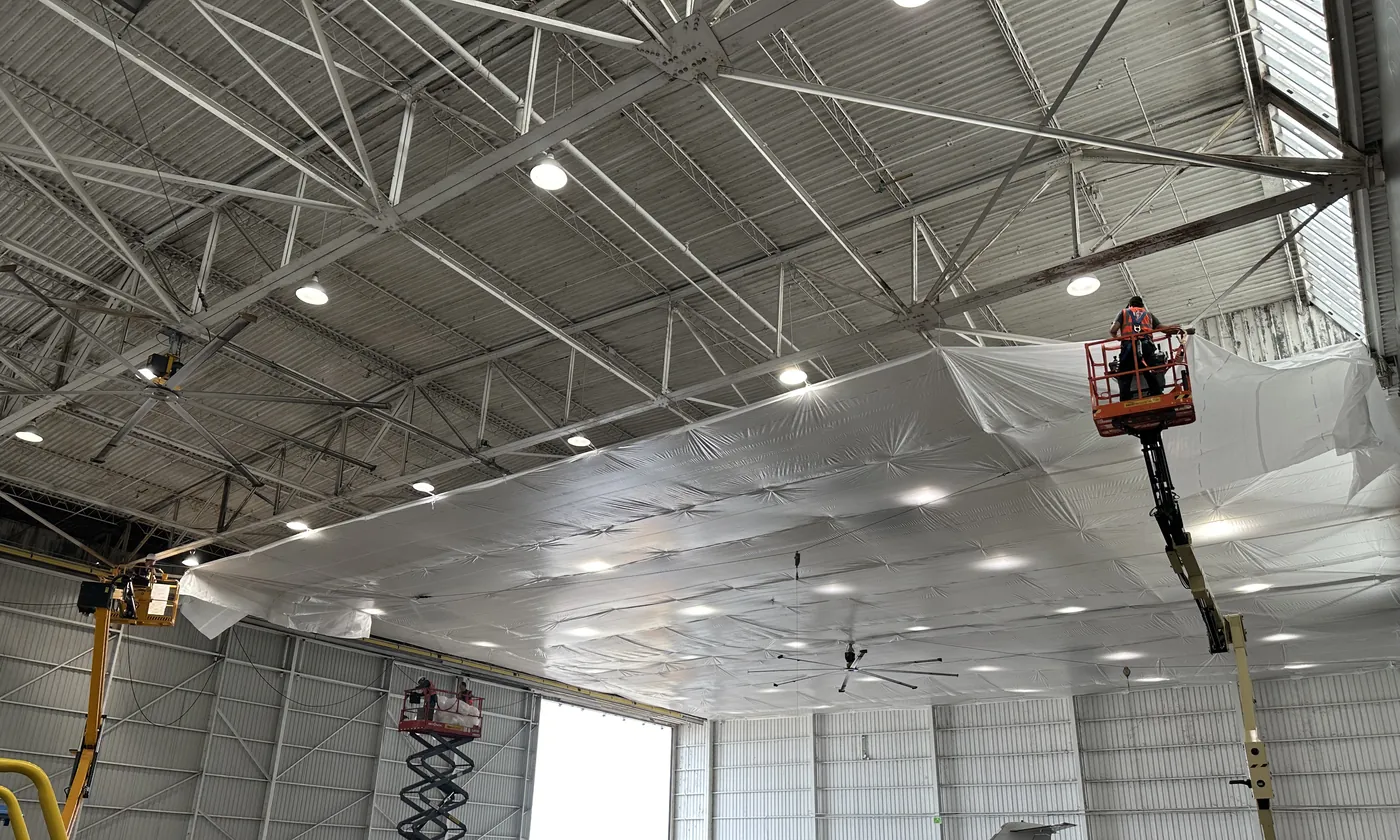 Inside a large hangar, plastic sheeting hung beneath the steel roof structure across much of the ceiling, with a worker on a boom lift at its edge and more lifts below