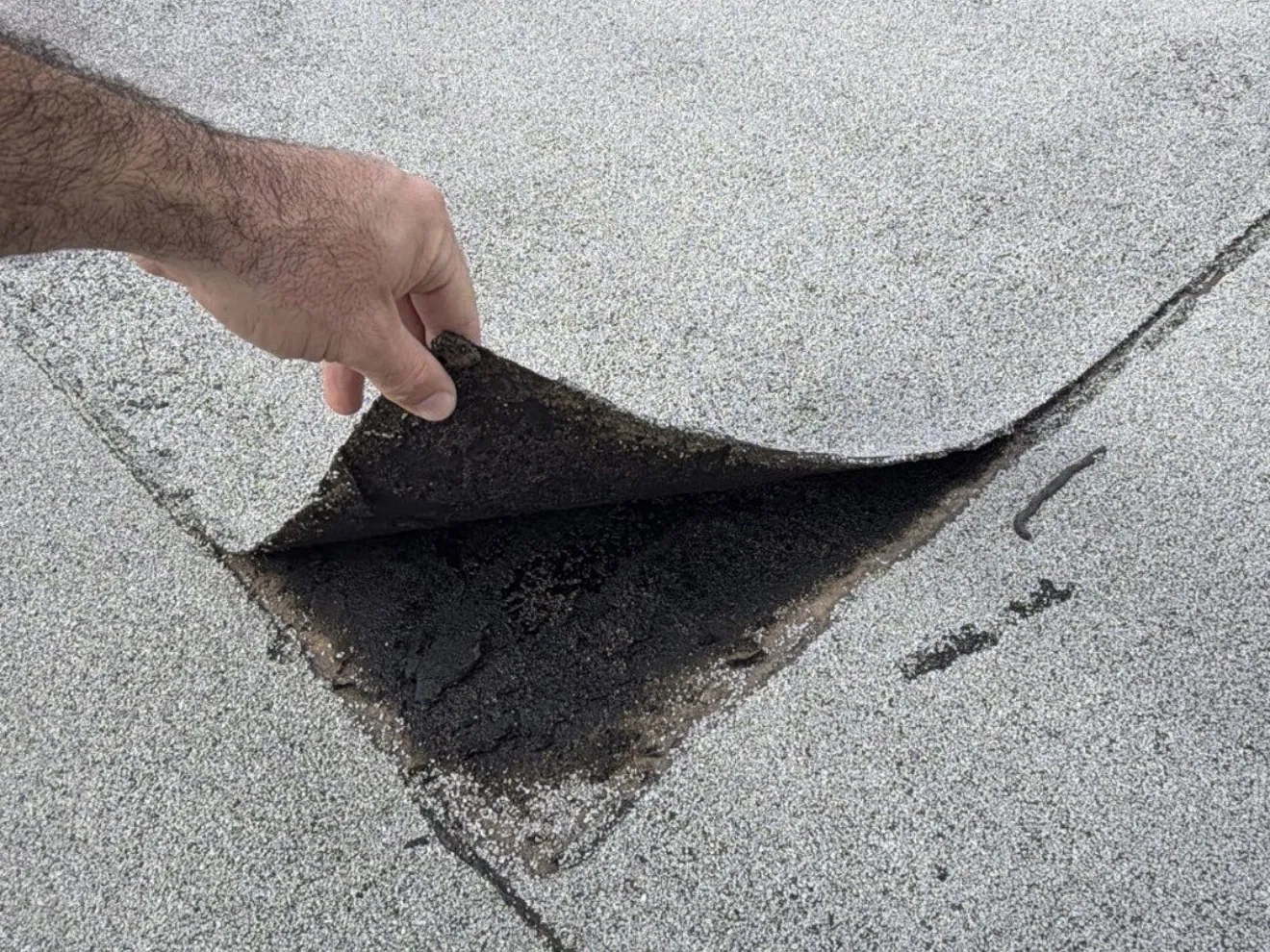 A hand lifting the loose corner of a granulated modified bitumen cap sheet where its seams meet, showing the black asphalt underneath