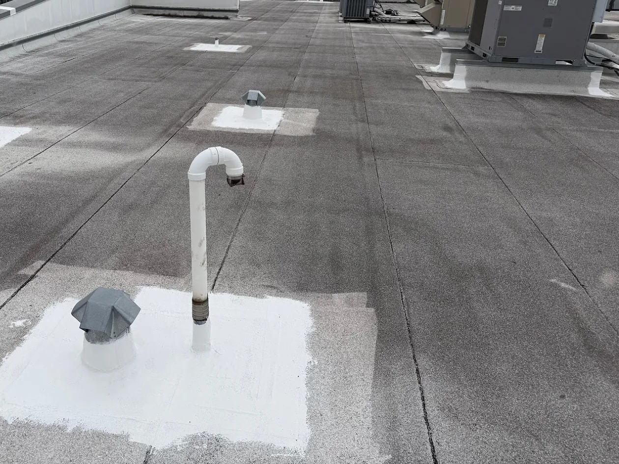 A granulated low-slope commercial roof with a hooked vent pipe and a capped vent sealed in a square of white coating in the foreground, more sealed vents further along the roof, and rooftop units on coated curbs at the back