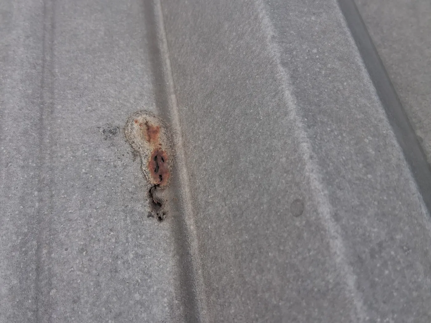 Close-up of a gray ribbed metal roof panel with a small patch of rust eaten through it beside a rib