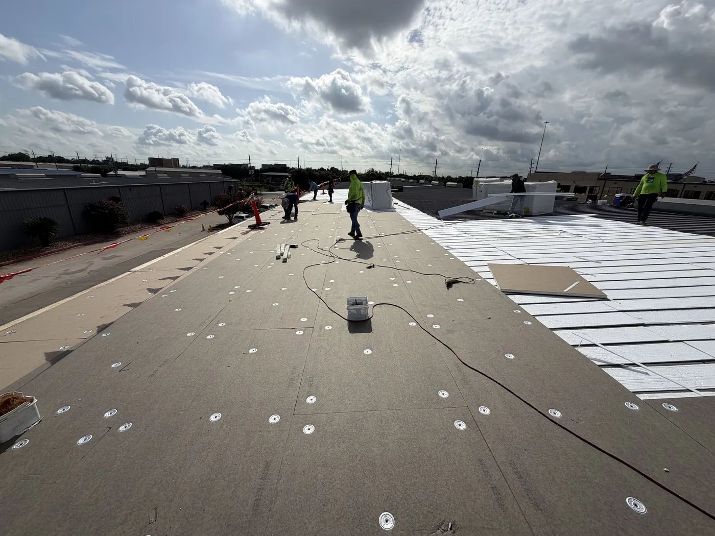 Insulation board fastened down with round plates across an existing metal roof, with bare metal panels and white foam strips between the ribs at right and crew working further along the roof