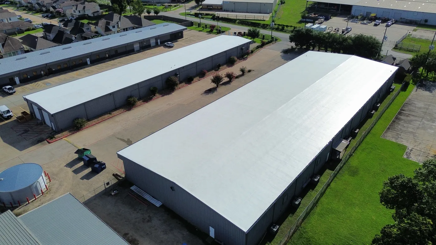 Aerial view of a long metal building with a smooth white roof, beside a second building with a white roof and an older metal roof beyond