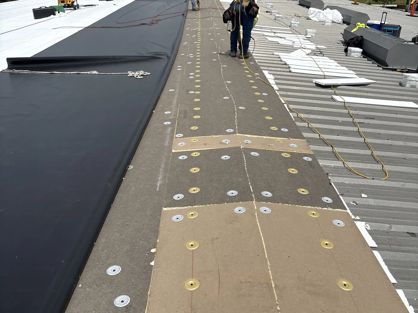 A metal roof retrofit in progress: bare ribbed metal panels with white foam strips and insulation on the right, board fastened down with round plates in the middle, and a dark membrane sheet rolled out on the left