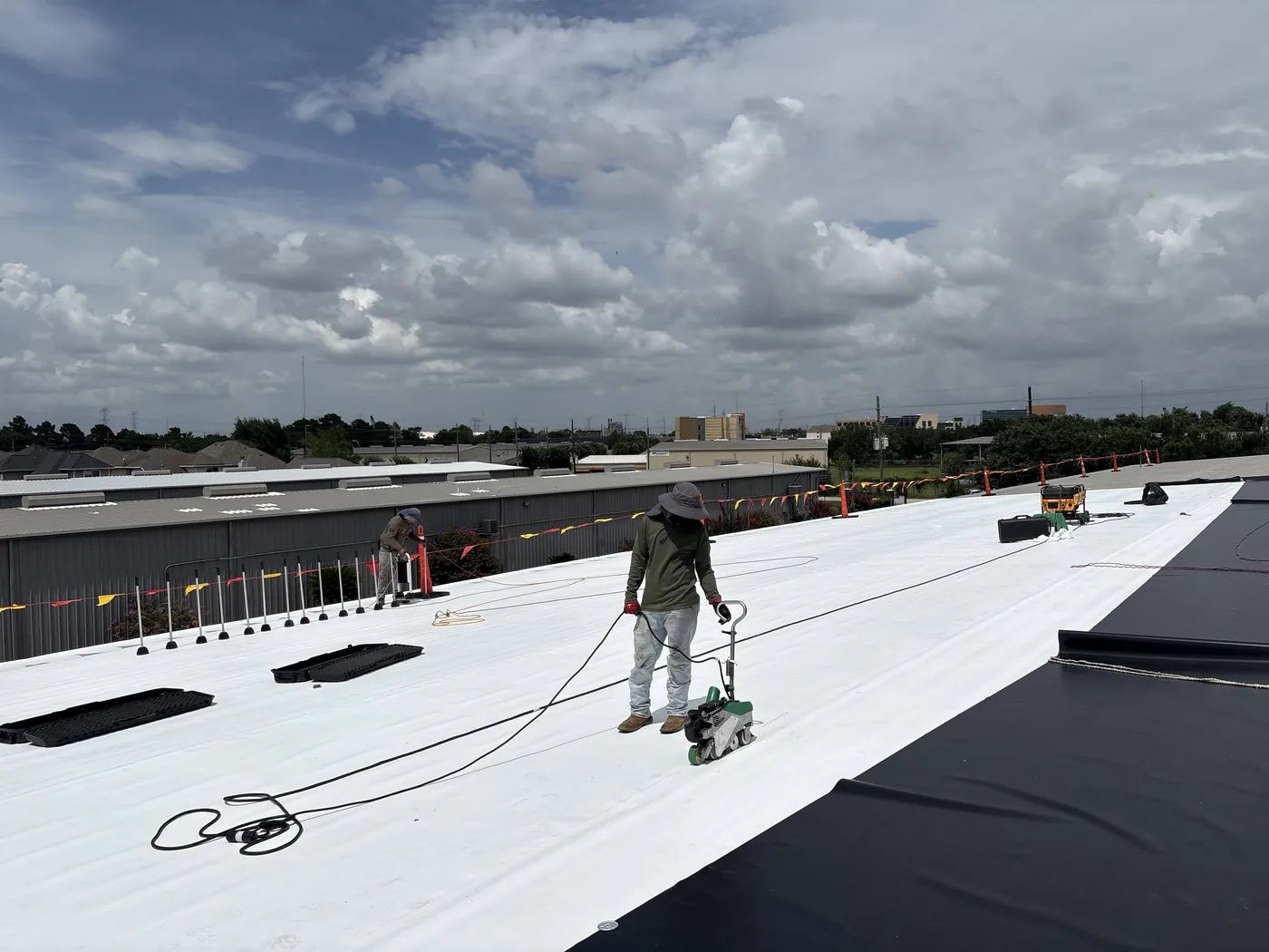 A roofer guiding an automatic hot-air welder along a seam in a white membrane roof, with a second roofer working near the edge, dark membrane sheets rolled out to the right and safety flags along the roof edge