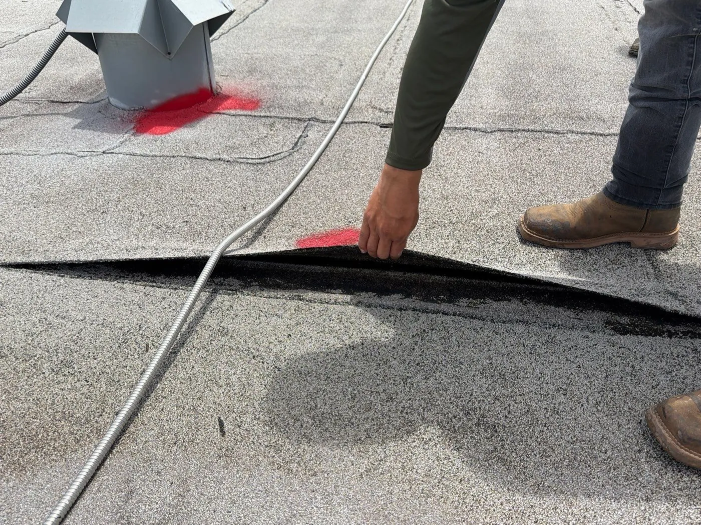 A hand lifting the edge of an open seam in a granulated modified bitumen roof, with red paint marks on the roof and at the base of a metal vent