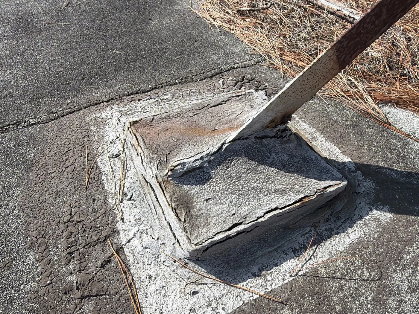 A weathered pitch pan around a rusted metal support on a granulated roof, its filler dried and cracked, with cracked roofing around its base and pine needles piled beside it