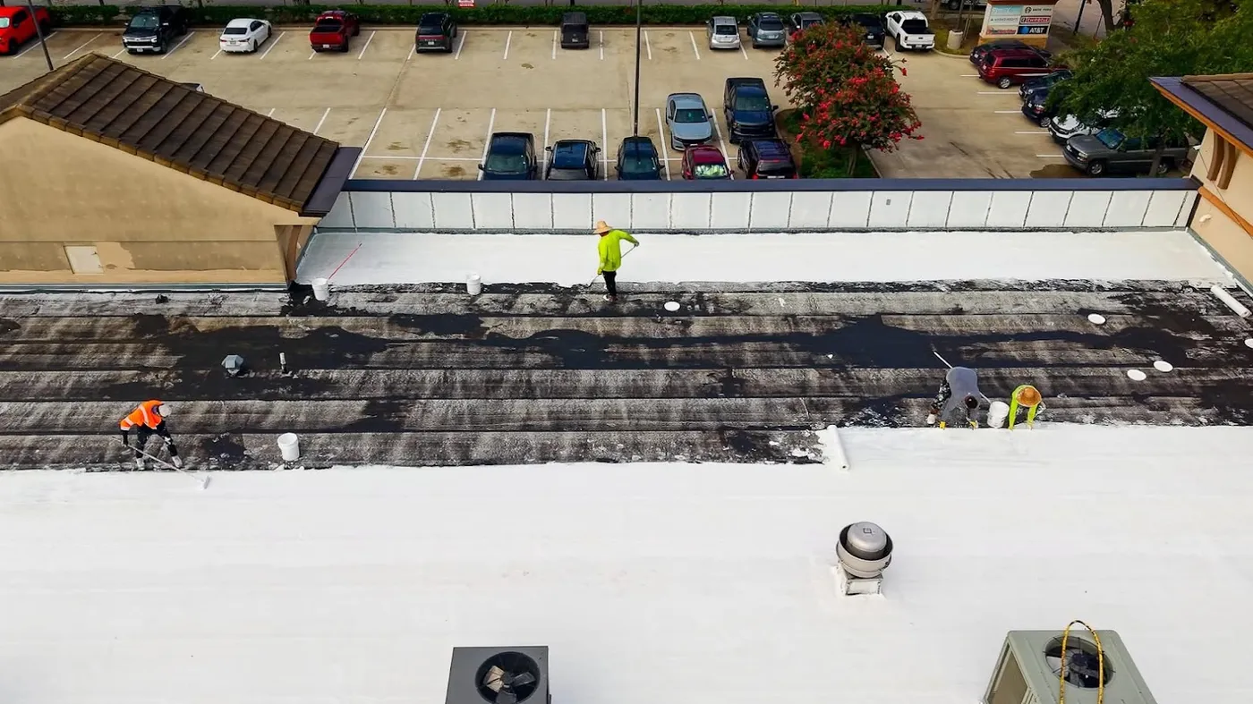 Aerial view of a small strip-center roof being coated in bands, with crews working across it and a partly filled parking lot beyond the parapet