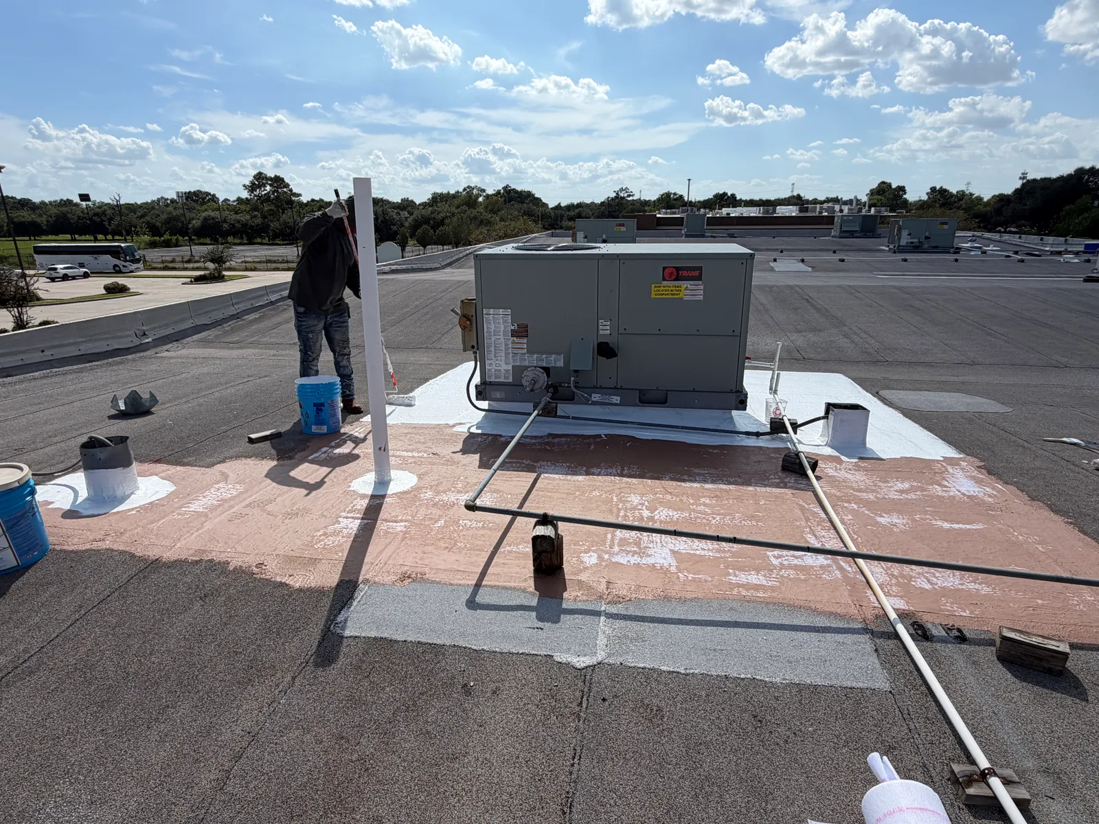 Packaged rooftop unit on a granulated roof ringed by a brown base coat with white top coat applied nearest the unit, one worker at the edge of the coated area and conduit supported on wooden blocks