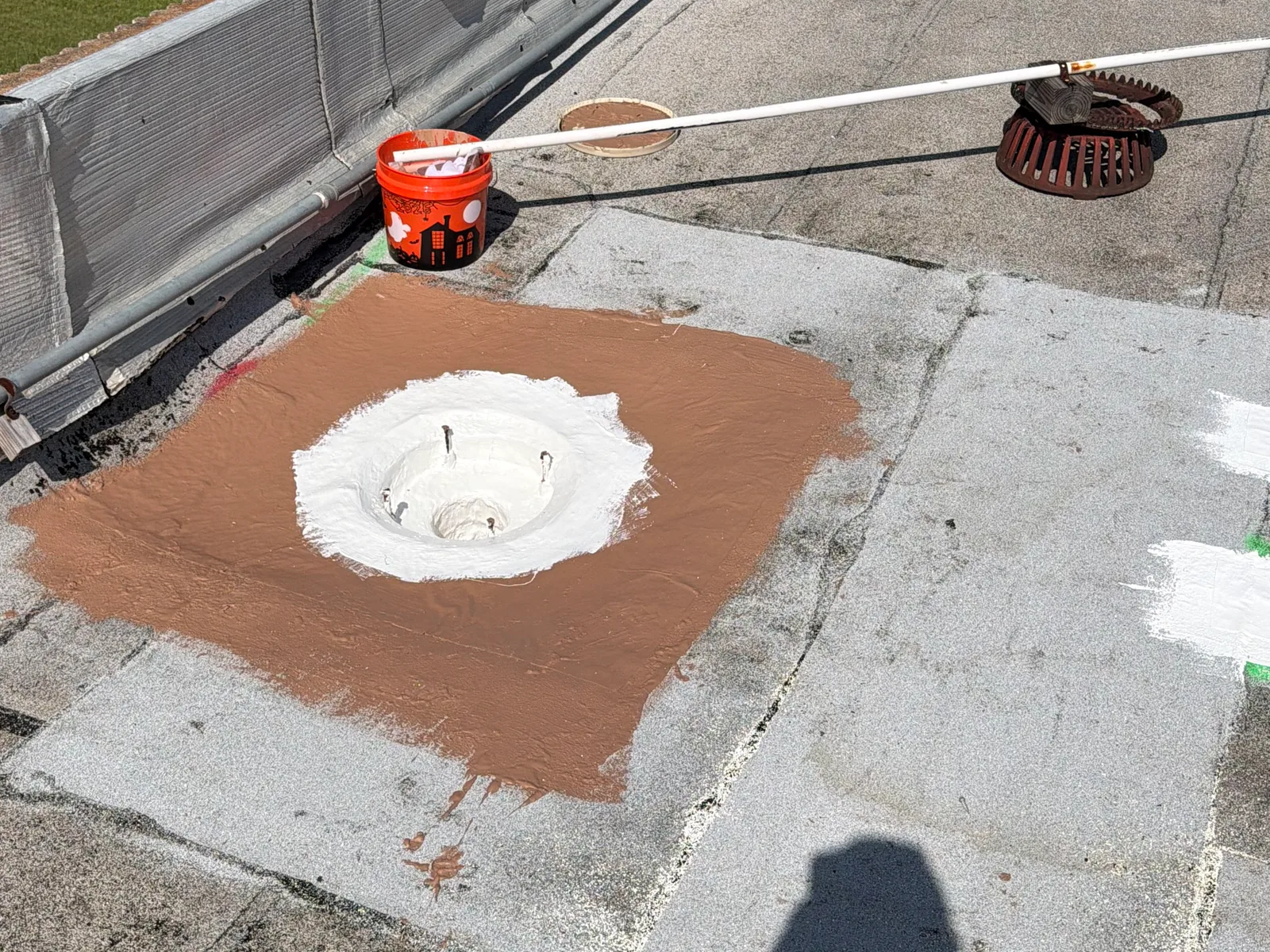 A roof drain with its strainer and clamping ring set aside and the studs exposed, the bowl coated white and a square of brown base coat around it on a granulated roof