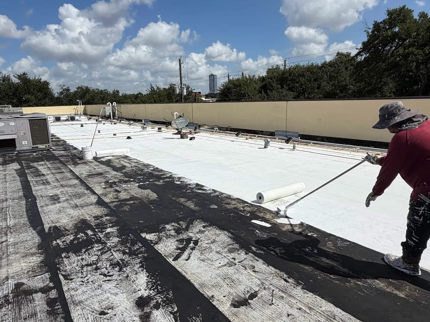 A roofer rolling white top coat over a darker layer on a low-slope roof, with a roll of reinforcing fabric on the deck