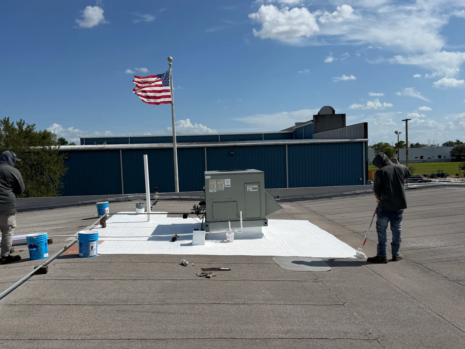 One roofer rolling white coating in a square around a rooftop unit and a vent pipe on a gray low-slope roof while a second stands by the buckets, with a blue metal building, a flagpole and a grassy lot beyond
