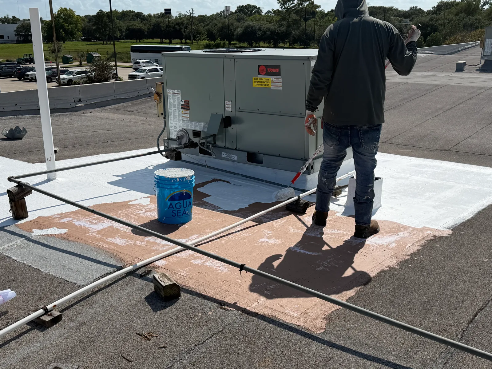A roofer rolling white top coat over a brown base coat around a packaged rooftop unit, with a gray pipe resting on wooden blocks and a bucket of coating beside it