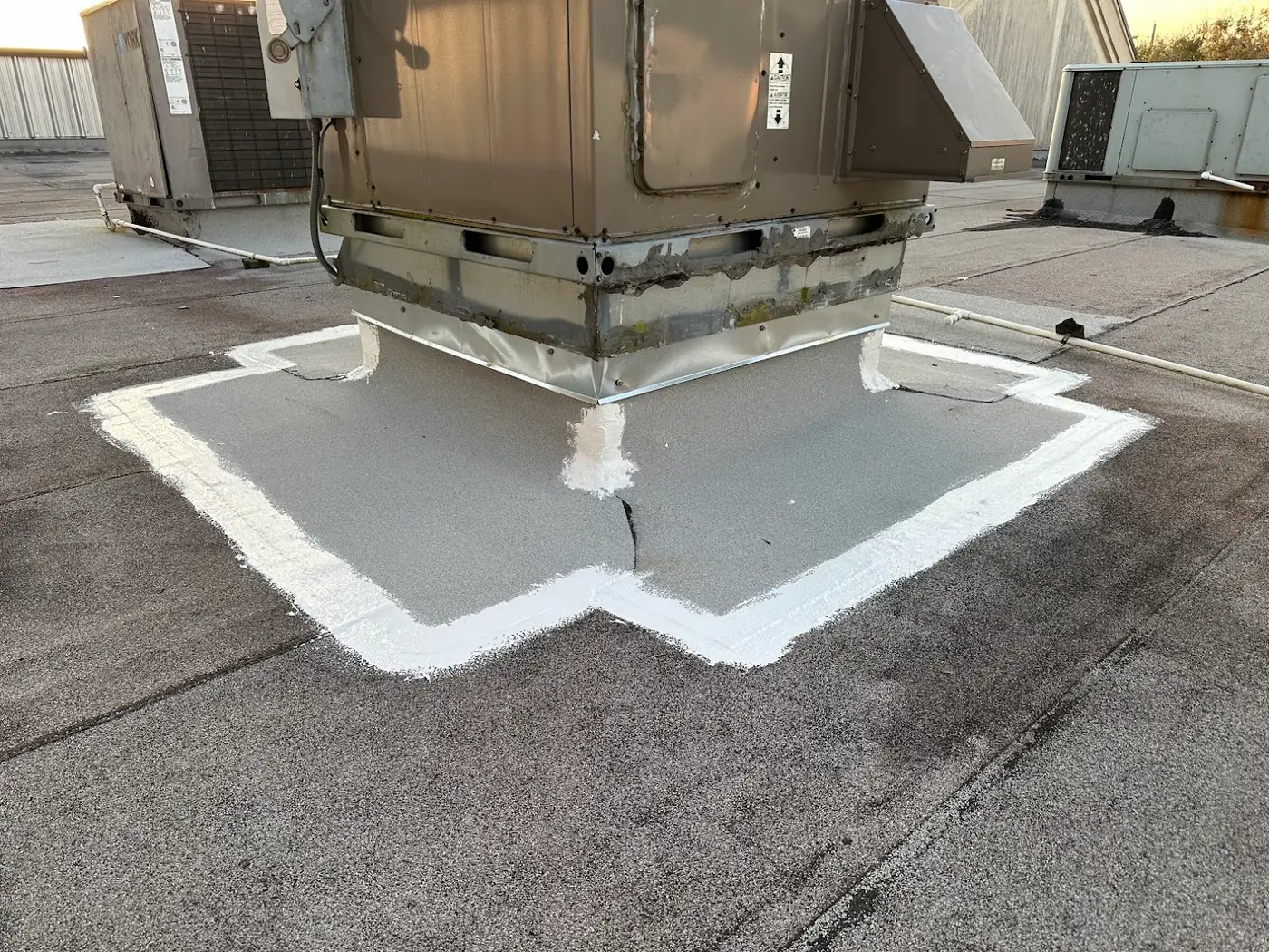 A packaged rooftop unit on a granulated low-slope roof, its curb base sealed in gray coating with a white border and a new galvanized skirt, with two more rooftop units and white pipes beyond