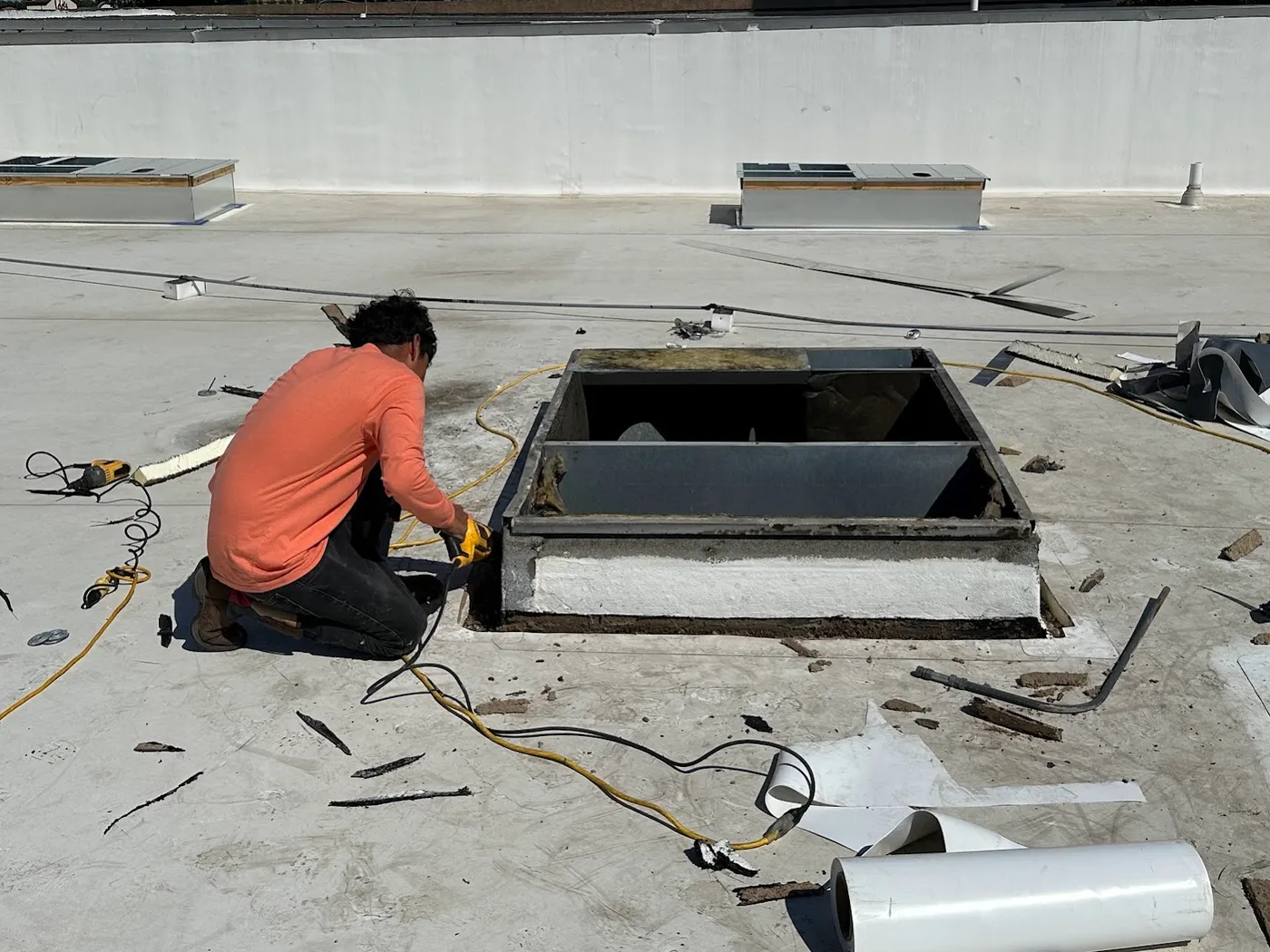 A roofer kneeling beside a large open curb on a white single-ply roof, cutting failed flashing away from its base, with a roll of white membrane in the foreground and two other rooftop curbs beyond
