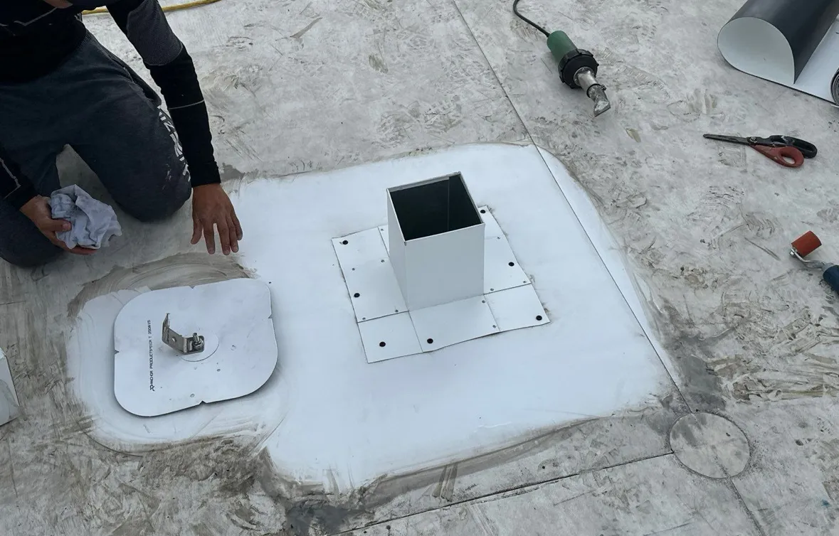 A roofer with a rag beside a new metal pitch pan fixed to white single-ply, the membrane around it wiped clean and the surrounding roof dirty and scuffed, with a hot-air welder lying unused nearby