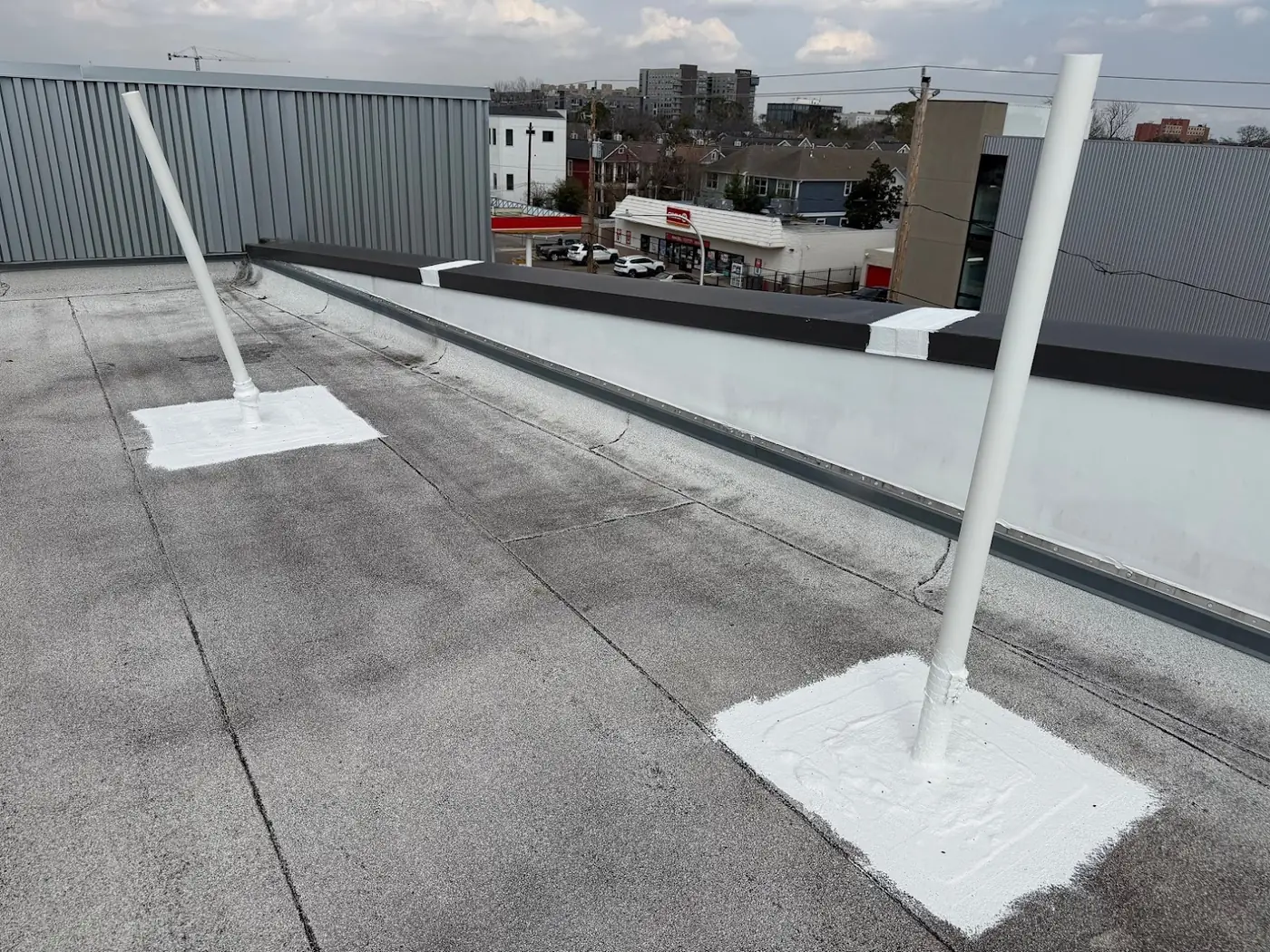 Sealed joints in a parapet coping and two white-sealed pipe bases on a granulated cap sheet, with a street and low buildings beyond the wall