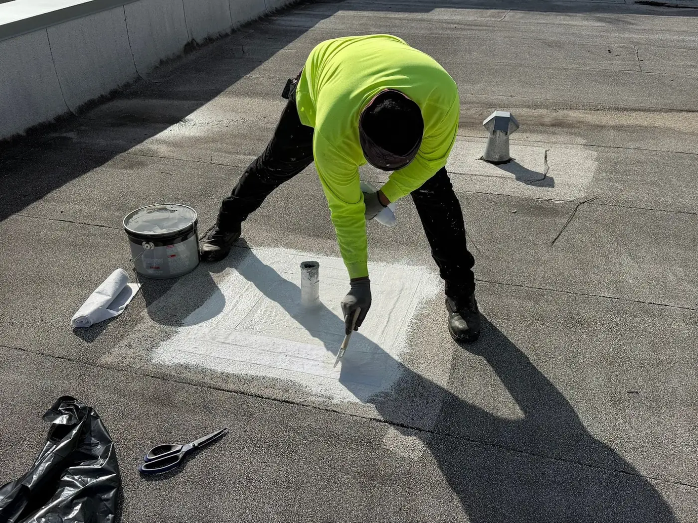 A roofer brushing white liquid flashing and fabric around a pipe penetration on a granulated low-slope roof, with a vent standing on the roof behind him