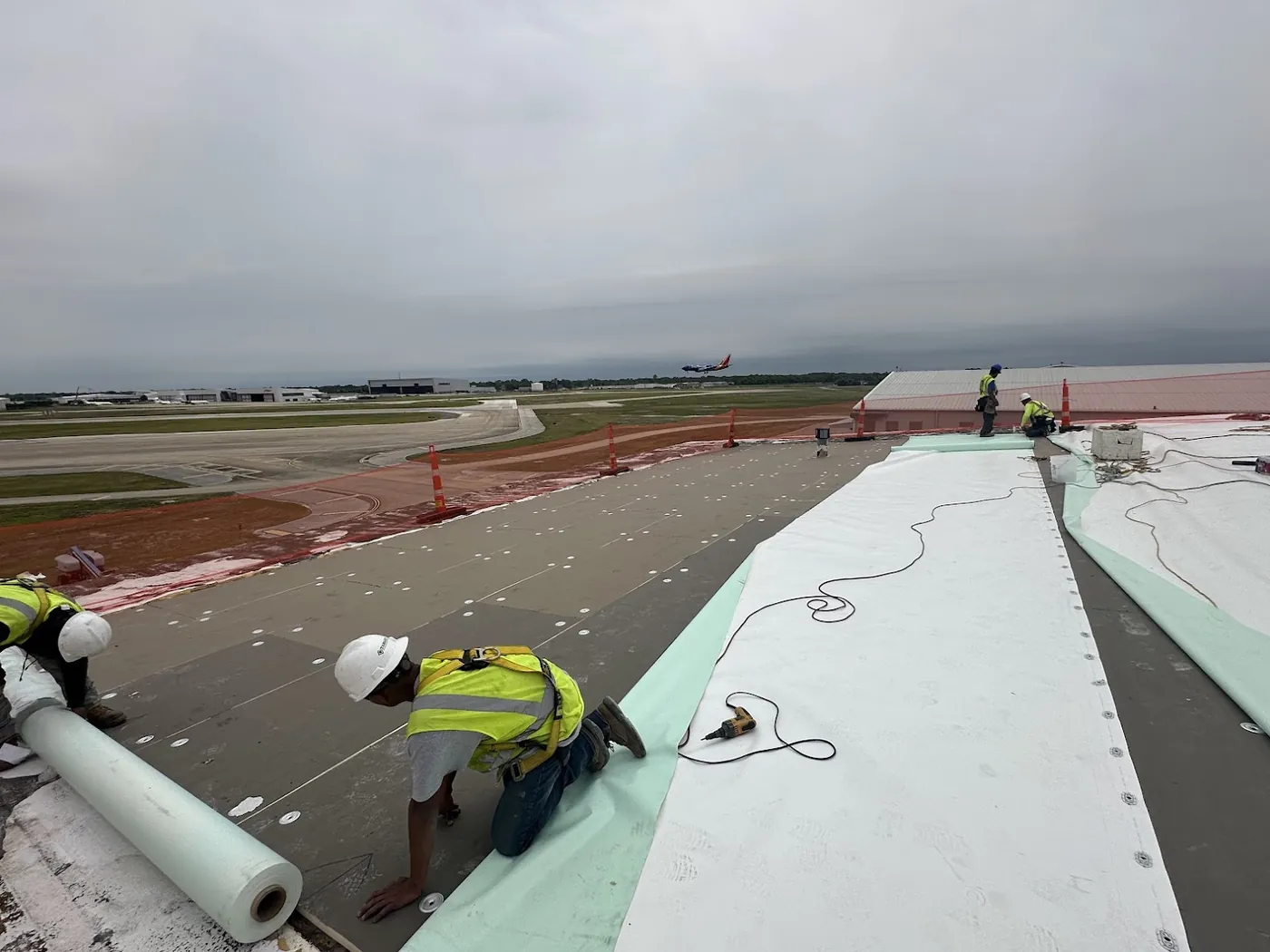 Roofers in hard hats and harnesses rolling out white single-ply membrane over gray insulation board fastened down with round metal plates on a low-slope roof