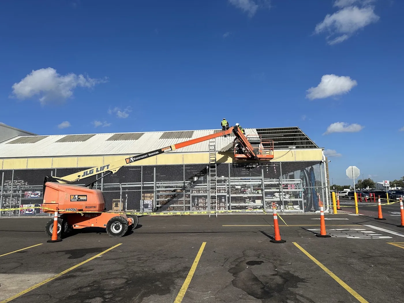 Two roofers on a metal panel roof beside a raised boom lift, with the far end of the roof stripped back to its framing and the parking lot below taped and coned off