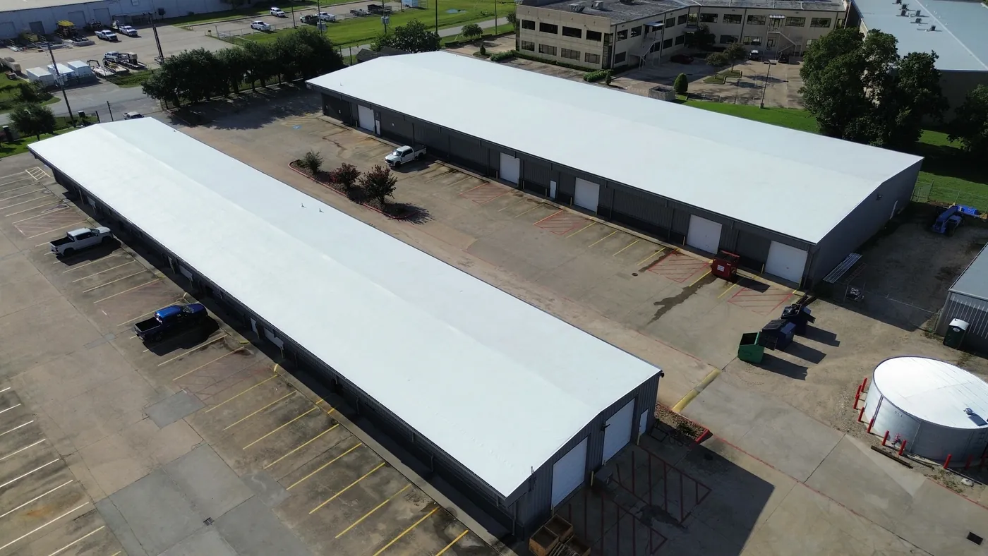 Aerial view of two long metal buildings with smooth white low-slope roofs, roll-up doors along their sides and a parking lot between them