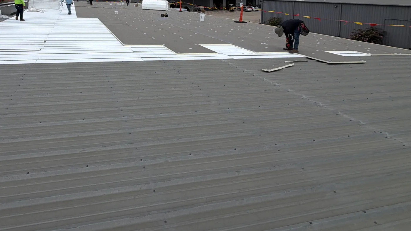 An existing ribbed metal roof with rows of exposed fastener heads in the foreground, white foam strips laid between the ribs beyond it, gray board fastened down with round plates over the strips, and a roofer bent over the board at right, with safety cones and flag line along the roof edge