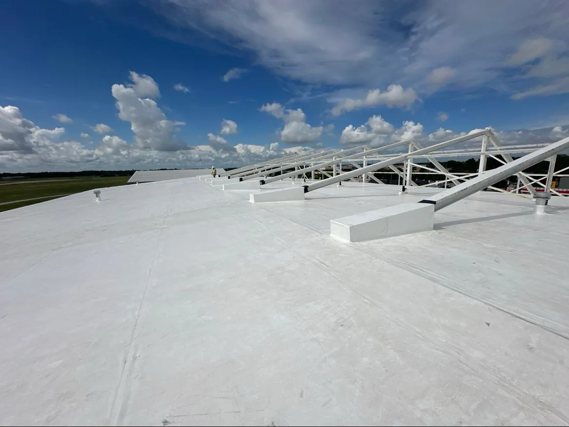A wide white membrane roof under a blue sky with scattered clouds, with seam lines running across it, pipe vents, a row of white steel frames standing on white blocks along the right side, and a worker in a hi-vis vest far along the roof